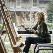 photo of a senior woman sitting in a chair next to a large floor-to-ceiling window, smiling and painting on an easel