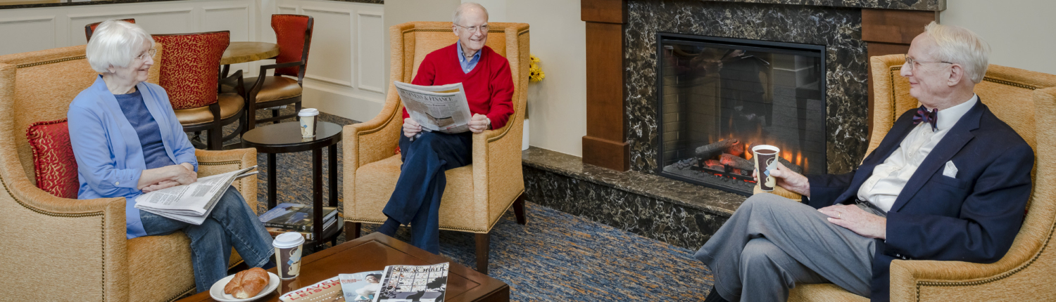 three seniors talking and reading the newspaper near a fireplace in one of Roland Park Place’s community lounge areas