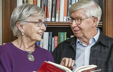 a senior man and woman looking at each other and smiling with a book open and standing in front of a bookcase