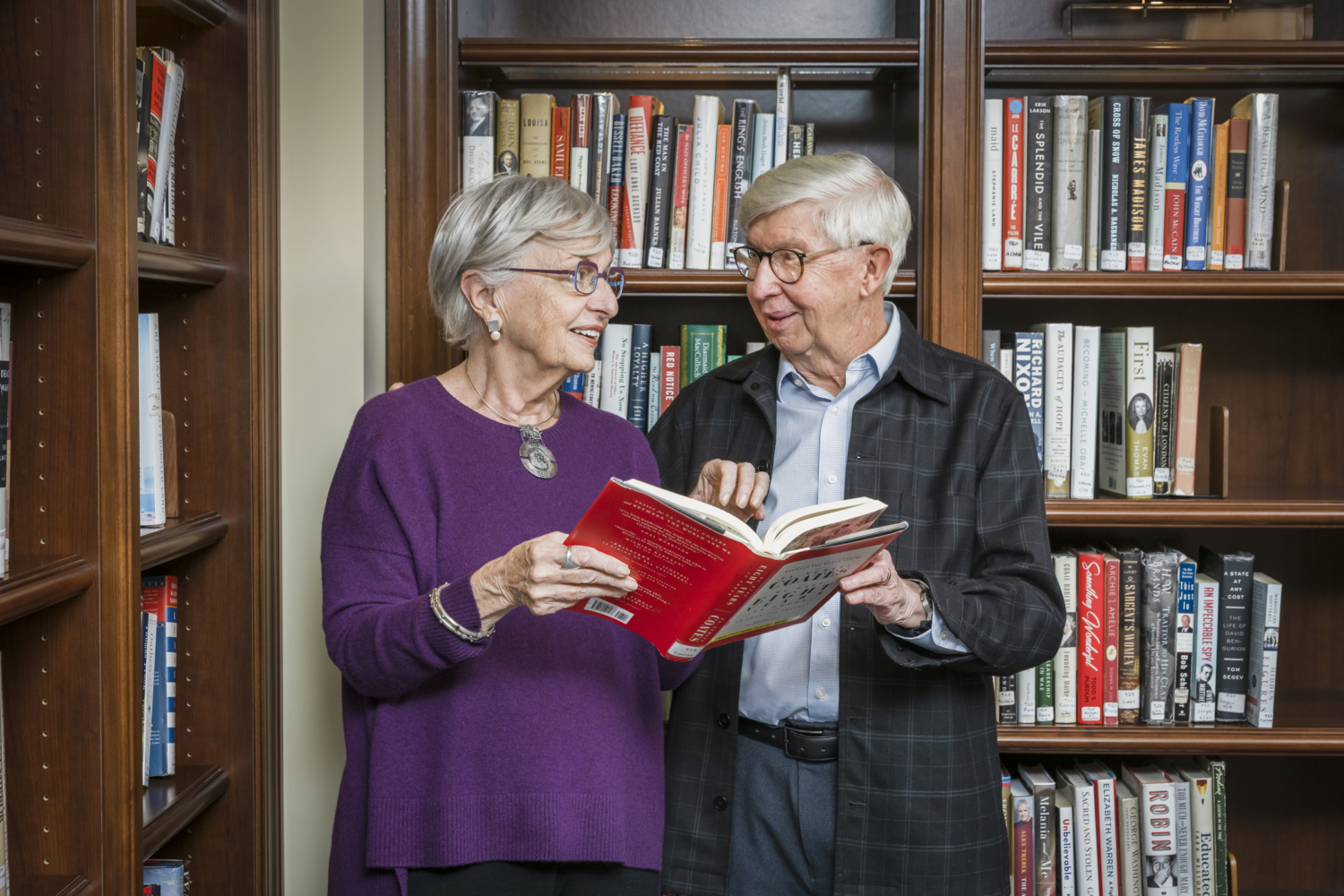 a senior couple standing in front of a bookcase reading a book together and talking