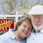 a senior couple standing in front of a "home for sale" sign with a SOLD sticker the sign. large home is in the background of the photo to illustrate that the couple is selling their home in order to downsize