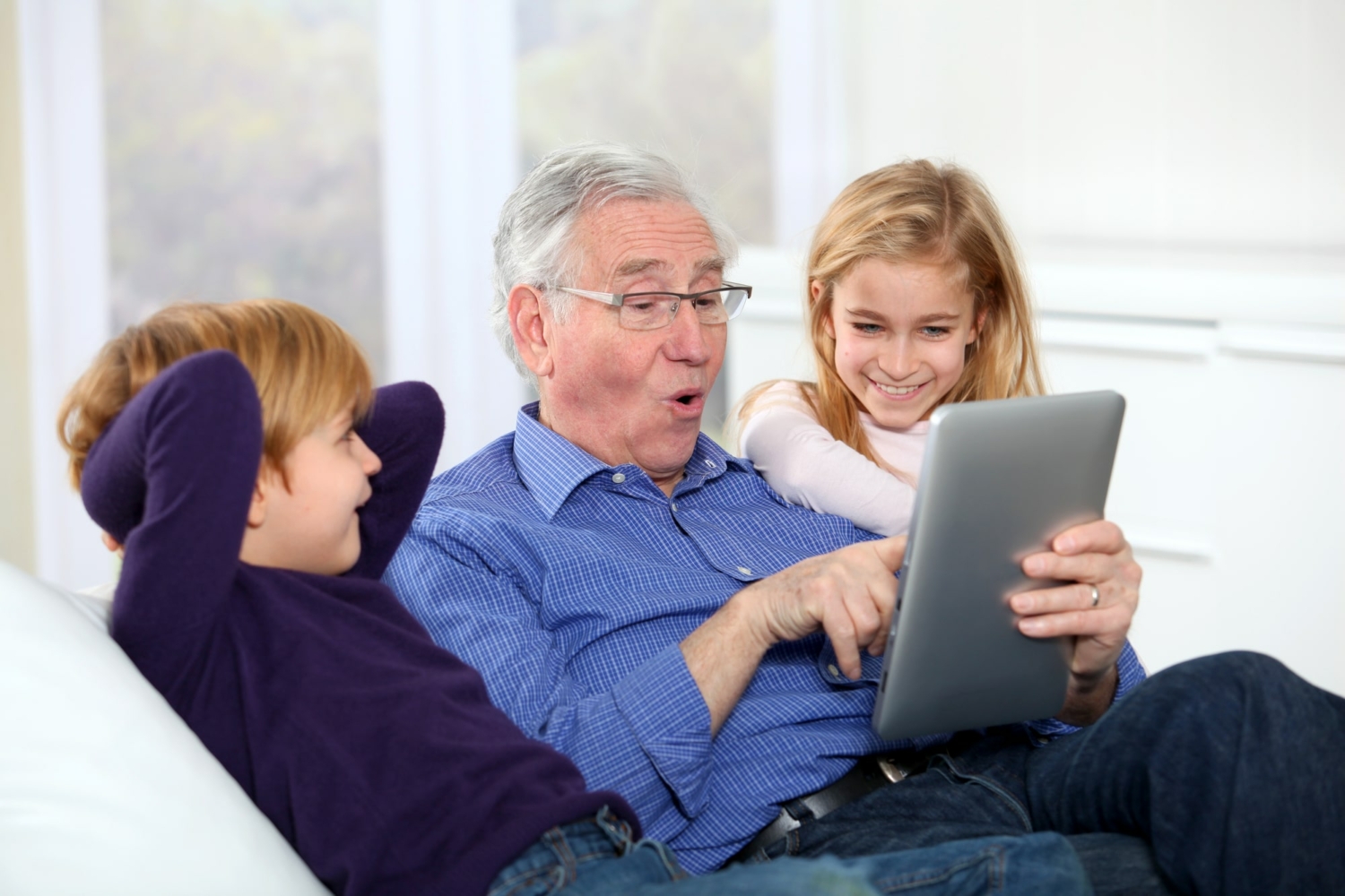 Senior man improving his tech skills on a tablet, sitting between two of his grandchildren on a couch.